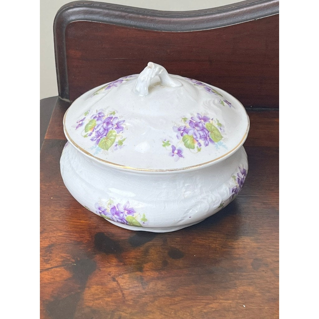An Antique Victorian Porcelain Wash Stand Set with Stand by Leland (late 19th c.) in white, detailed with purple and green floral motifs, rests on a polished walnut stand; a dark wood chair appears in the background.