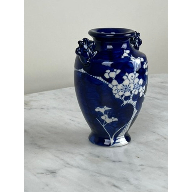 The Vintage Japanese Blue and White Floral Bud Vase, featuring ornate handles and white floral patterns, is displayed on a light marble surface against a plain background.