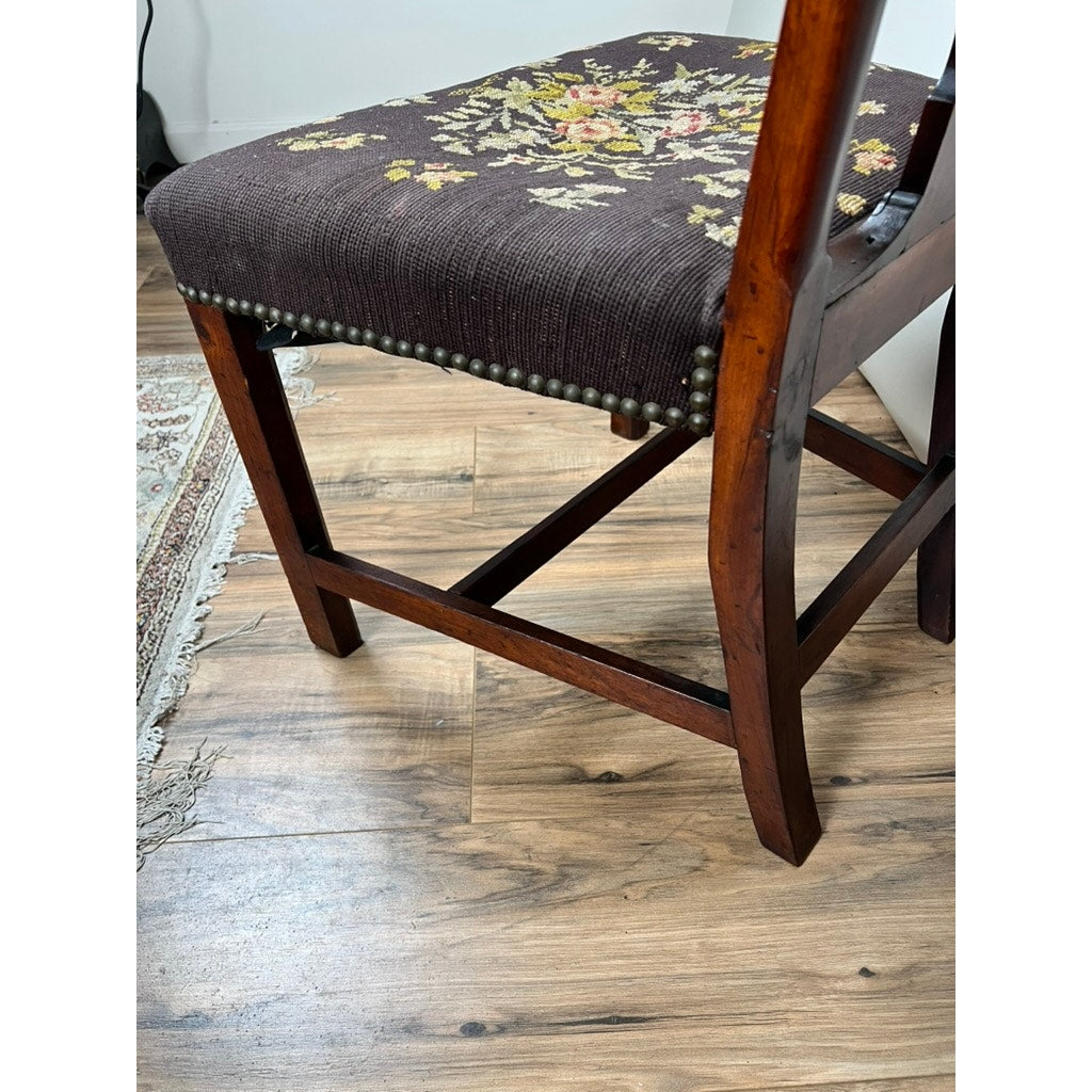 An Antique Chippendale Side Chair with a mid-19th-century needlepoint seat rests on a wood floor near the corner of a patterned rug, its edge accented with metal studs. Brand: None/Unknown.
