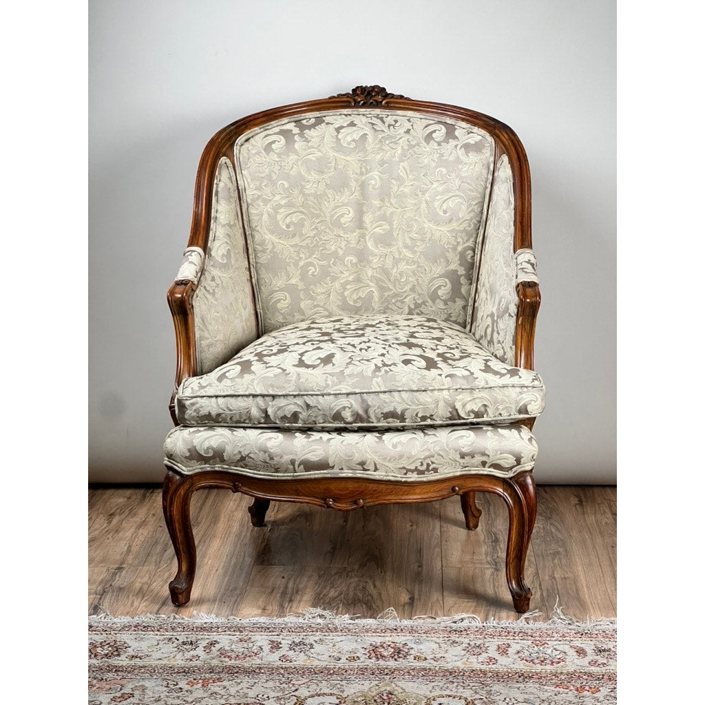 An Antique French Louis XV Rococo Style Bergère Arm Chair, c. mid 19th century, with ornate wood frame and newer damask upholstery, is shown on a wooden floor with part of a patterned rug in the foreground.