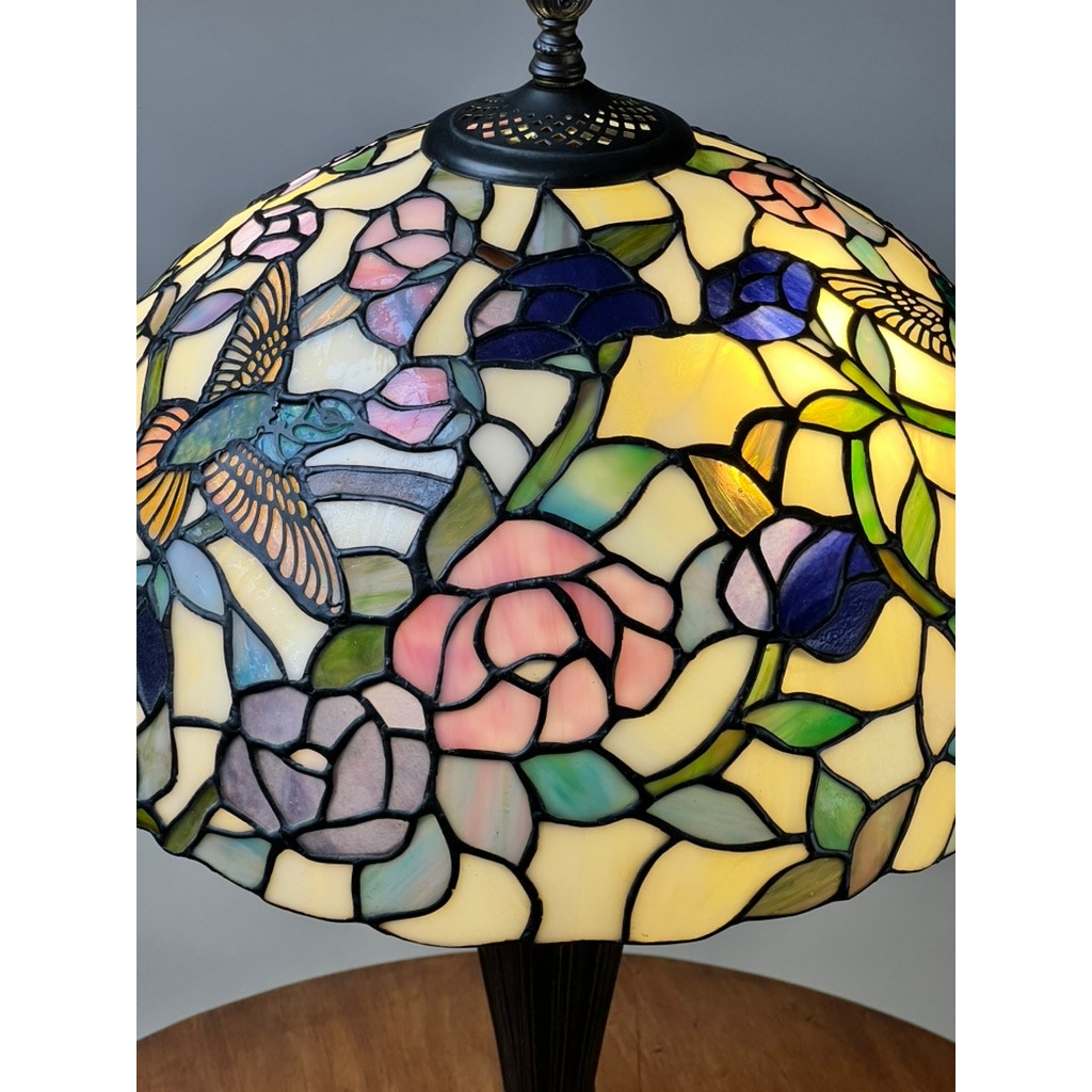 A close-up of a Vintage Tiffany Style Floral Stained Glass Table Lamp with colorful Art Nouveau flower and hummingbird designs in pink, purple, blue, and green shades, beautifully illuminated from within. Brand is unknown.