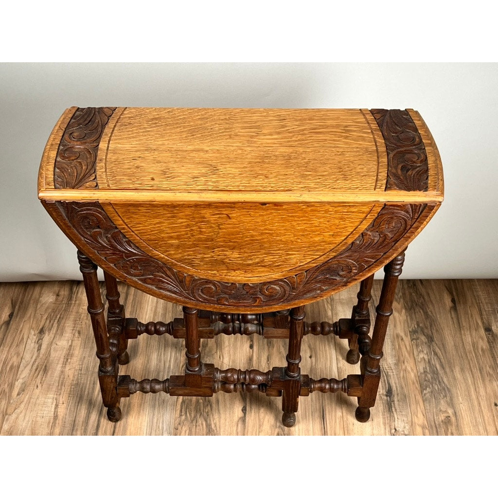 An Antique English Oak Carved Gateleg Occasional Table c. Late 19th Century, featuring detailed carved edges and turned legs, stands on a wood-patterned floor against a plain light-colored wall. Brand: None/Unknown.