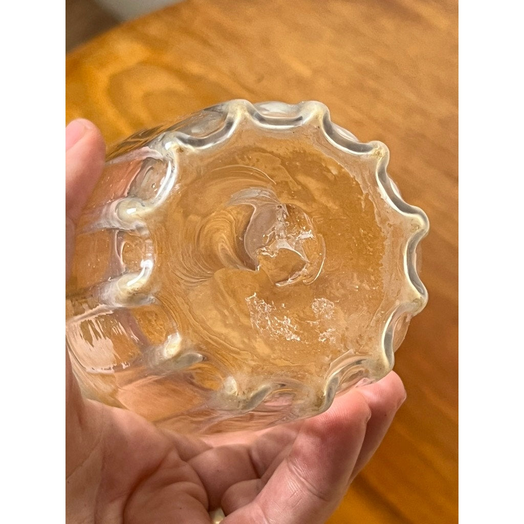 A hand holds the Attributed Boston & Sandwich Wheel Engraved Mold-Blown Glass Table Vase, circa 1845–1860, featuring a clear body with a wavy, scalloped bottom edge reminiscent of Victorian art glass. Textured patterns and small bubbles are visible in the glass, set against a wooden background.