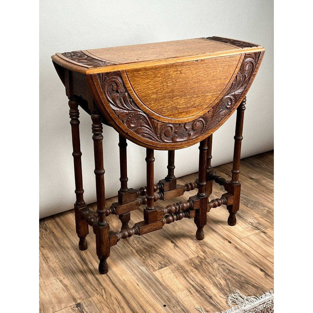 An Antique English Oak Carved Gateleg Occasional Table from the late 19th century, featuring ornate floral details and turned legs with one leaf extended, stands on a wooden floor against a plain light-colored wall.