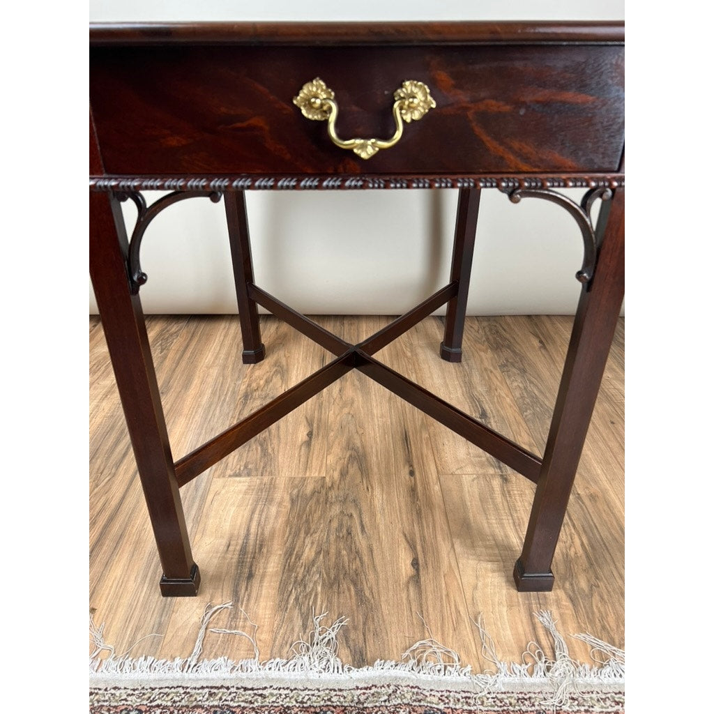 A close-up of the Stickley Vintage Federal Style X Stretcher One Drawer End Table, featuring a decorative brass handle, carved details, and crossed stretcher legs, standing on a wood floor beside a fringed rug.