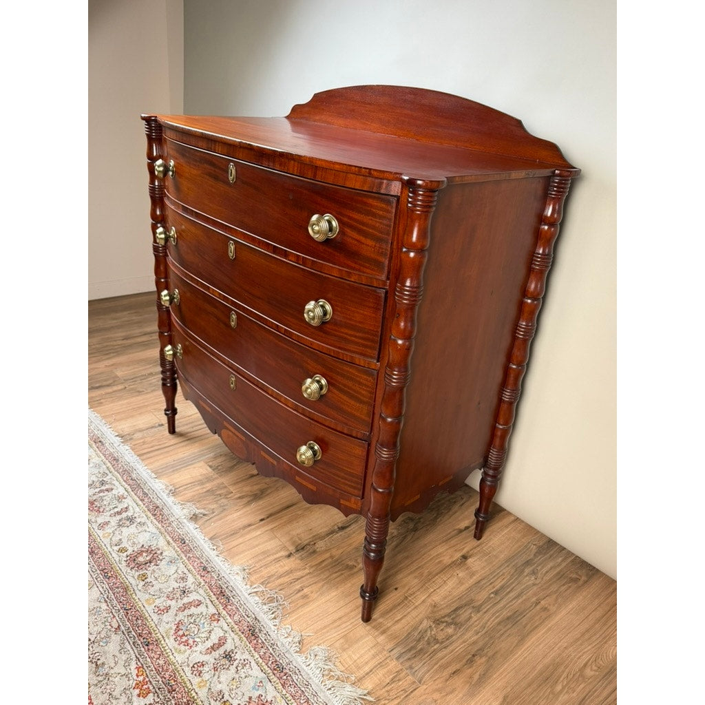 Early 19th Century New England Federal Mahogany Bow-Front Chest of Drawers with Backsplash, c. 1820–1840—this refined antique features four elegantly curved drawers, brass handles, and decorative turned side columns, beautifully displayed on a wooden floor beside a patterned rug and embodying the classic Federal style.