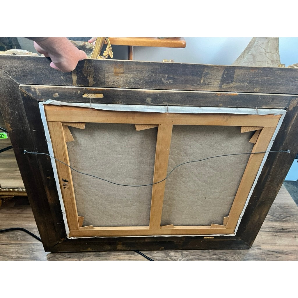 The image shows the back of a Vintage Guy Cambier Belgian Romantic Landscape Oil Painting with Horsemen (c. 1960–1980), featuring its wooden frame, canvas, and hanging wire. A hand holds the top edge as it rests on a wooden floor near some furniture.