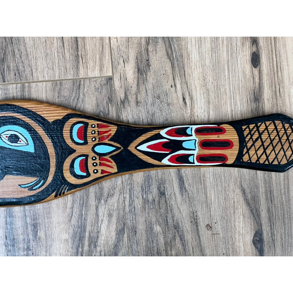 A Vintage Inuit carved red cedar paddle from Sitka, Alaska, signed illegibly, featuring bold black, red, and turquoise Indigenous patterns and an animal face, lies on a wood floor.