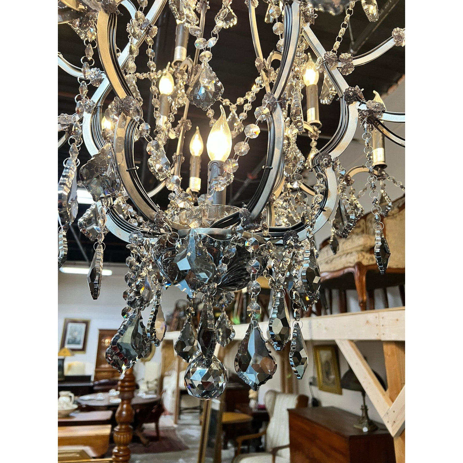A close-up of the Large Vintage Smoke Crystal 24 Light Chandelier with hanging glass prisms and lit bulbs, showcased in a room adorned with vintage wooden furniture and framed artwork in the background.