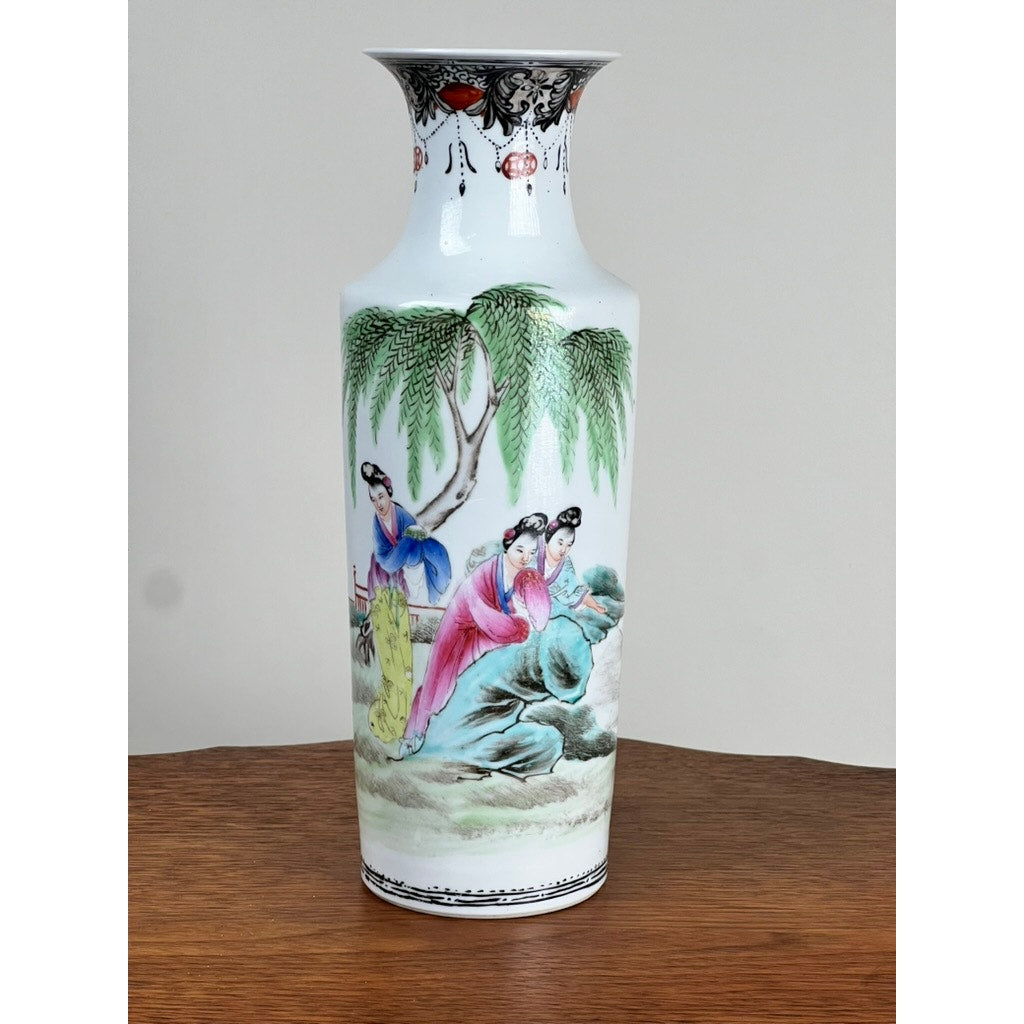An Antique Qing Dynasty Famille Rose 12” Vase with Qianlong mark, featuring a narrow neck and painted with three women in colorful traditional attire under a willow tree on a plain background, is shown resting on a wooden surface.