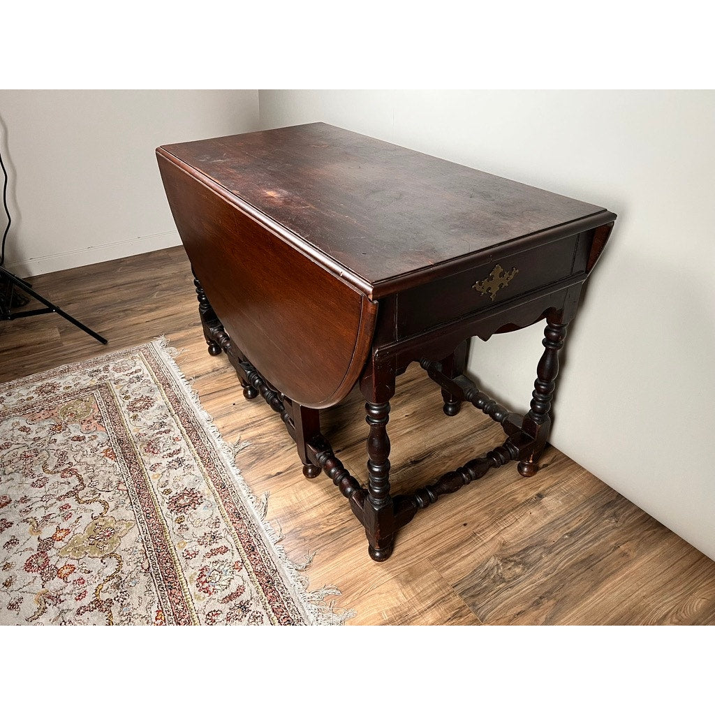 An Antique English William & Mary Walnut One Drawer Gateleg Dining Table, c. Early 18th Century, sits on a wooden floor beside a patterned rug in a white-walled room.