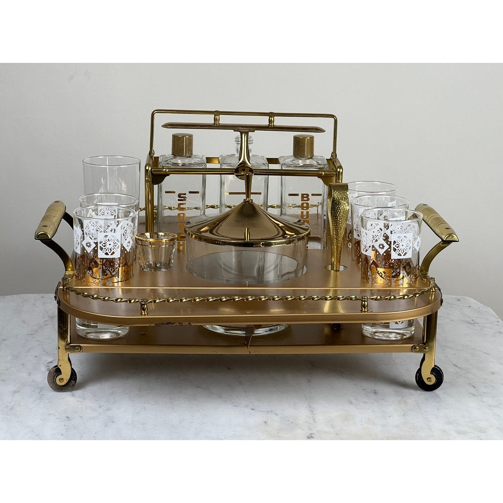 The Vintage Mid Century Modern Bar Caddy c. 1960s features brass, glass, handles, decanters, gold-trimmed glasses, a central ice bucket, and cocktail accessories, standing out against a plain white background. Brand: unknown.