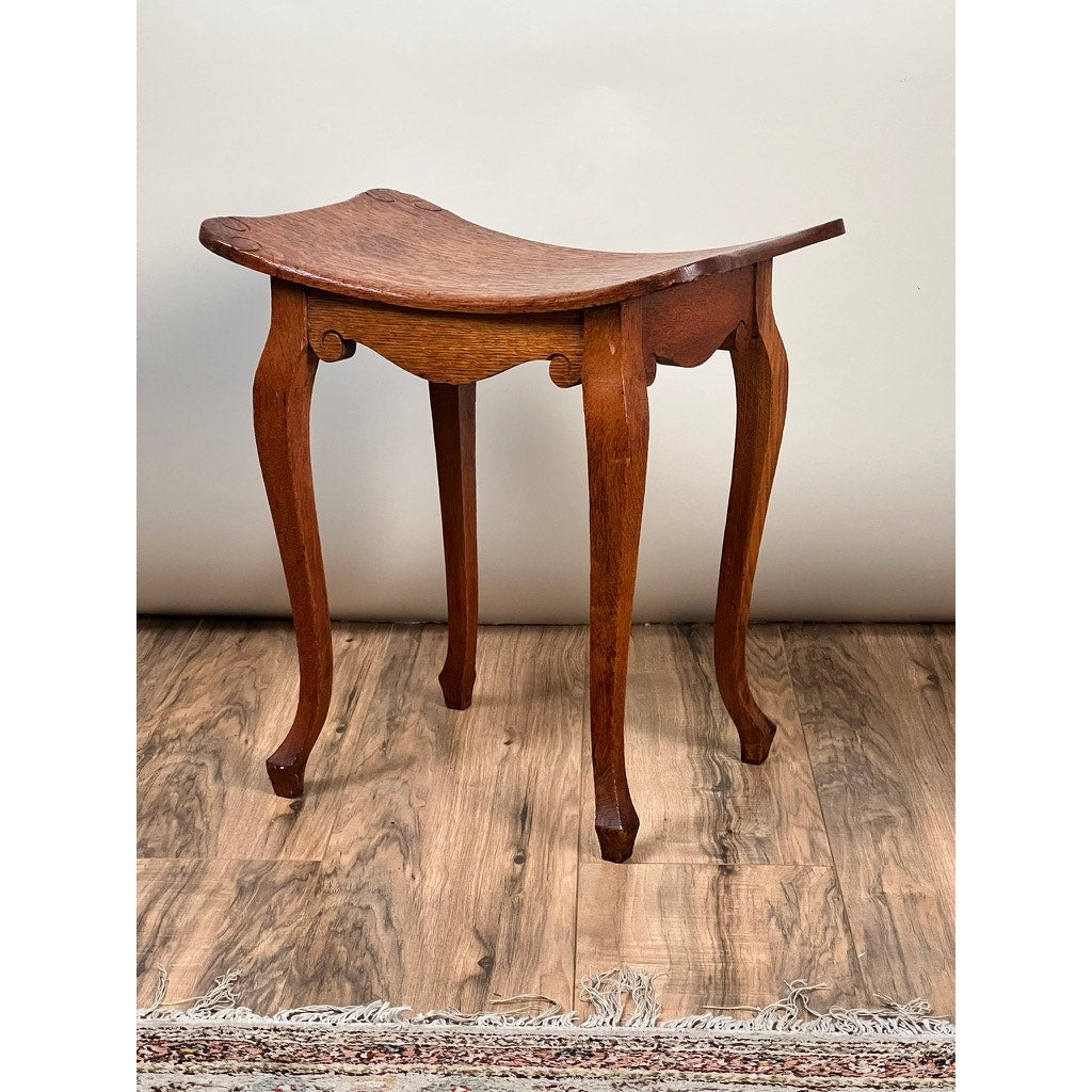 An Antique Arts and Crafts Saddle Shaped Footstool c. 1900 with cabriole legs rests on a wood floor beside a fringed rug and light wall, showcasing timeless charm and classic design. Brand: None/Unknown.