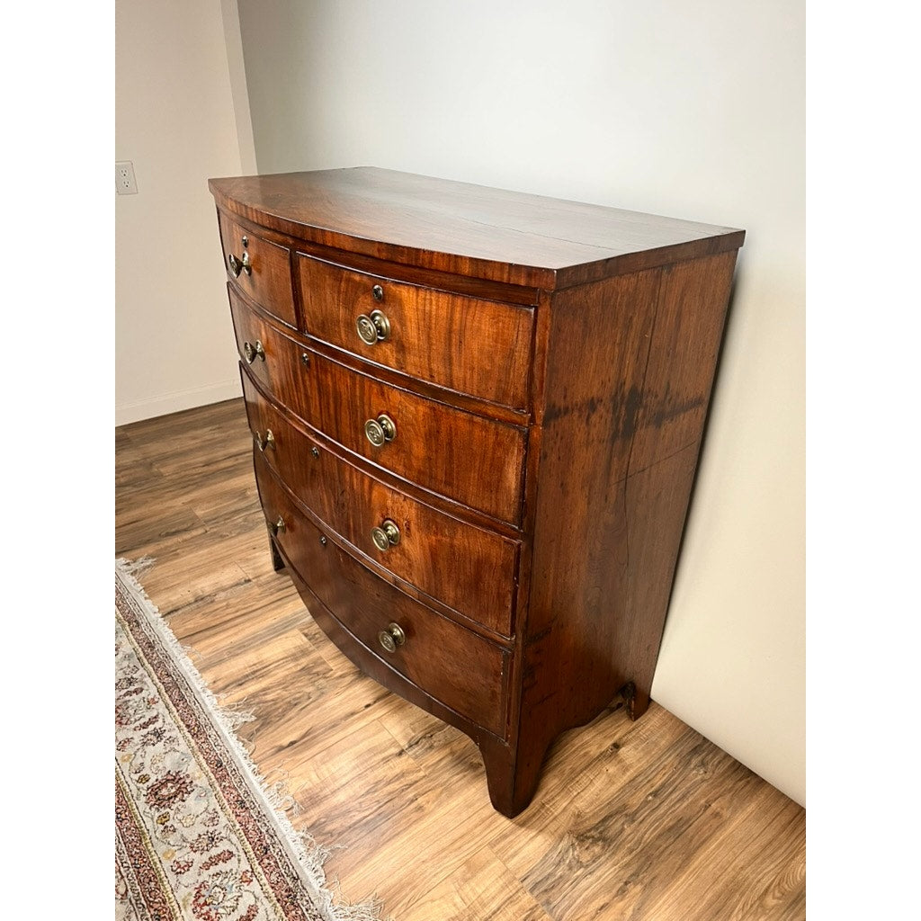 An Antique George III Mahogany Bowfront Chest on French Feet, c. Late 18th Century, with a polished finish and brass handles, stands on a wood floor near a beige wall and bordered rug.