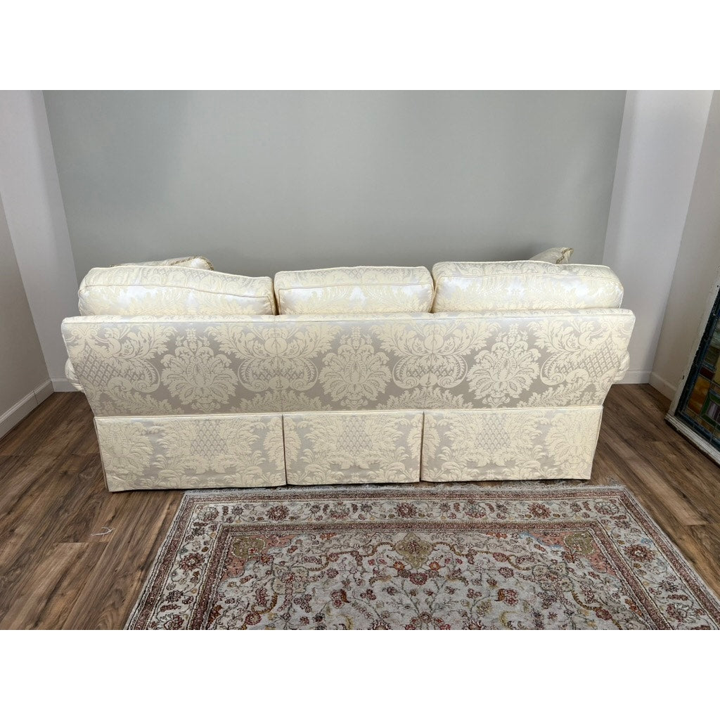 A Vintage Custom Ivory Upholstered Sofa by Stickley with three back cushions is placed against a light gray wall on a wooden floor, partially on an ornate floral area rug.