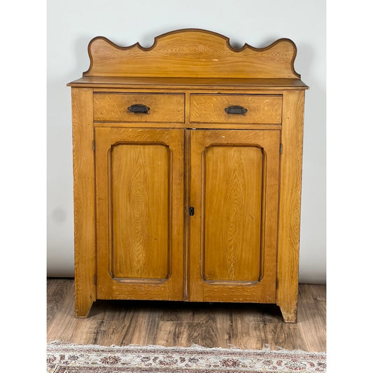 The Antique Victorian Grain Painted Country Jelly Cupboard c. 1870 by Pearl City Antiques features two drawers on top, two paneled doors below, decorative top trim, and metal handles. Resting on a wooden floor with a glimpse of a patterned rug, this piece offers an inviting example of 19th-century American craftsmanship.