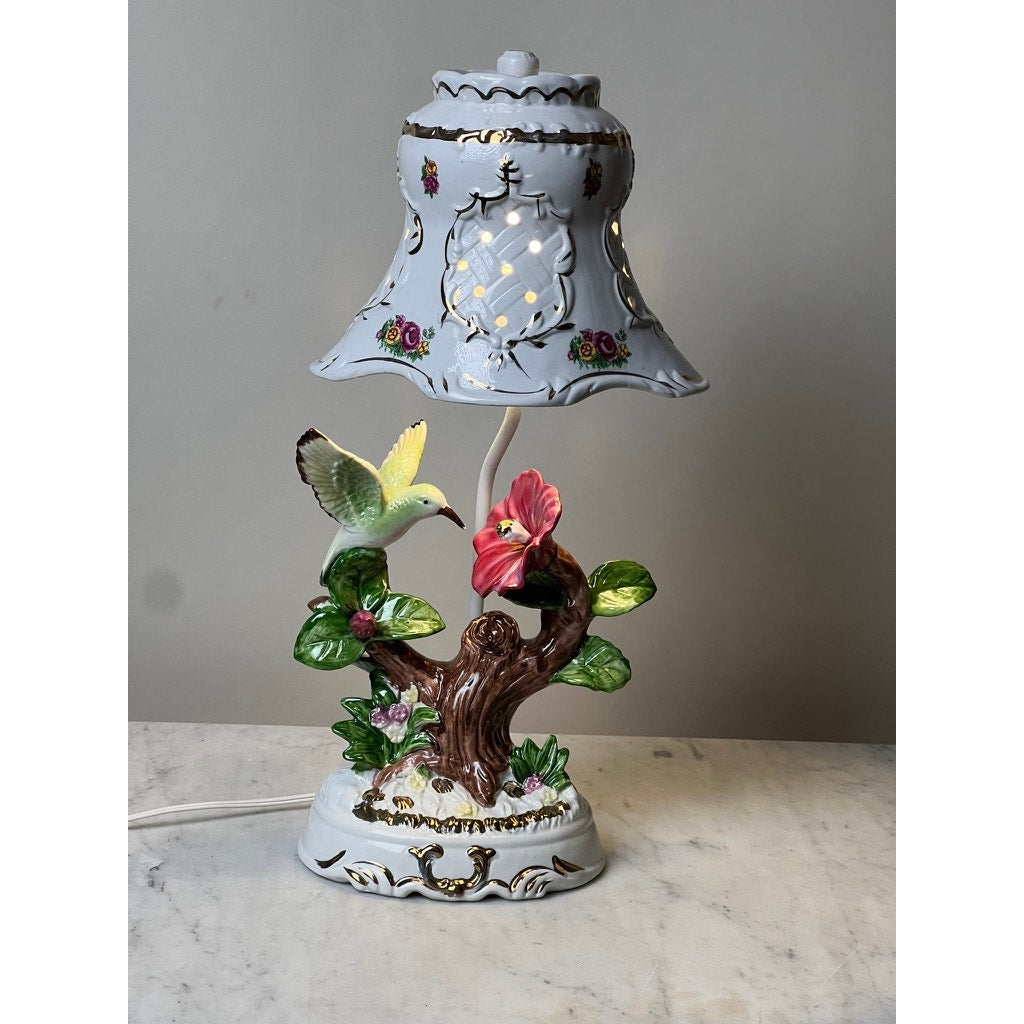 The Vintage Lena Liu Porcelain Hummingbird Accent Lamp features a colorful hummingbird by a pink flower on a tree trunk with green leaves and gold accents, elegantly displayed on a marble surface against a plain background.