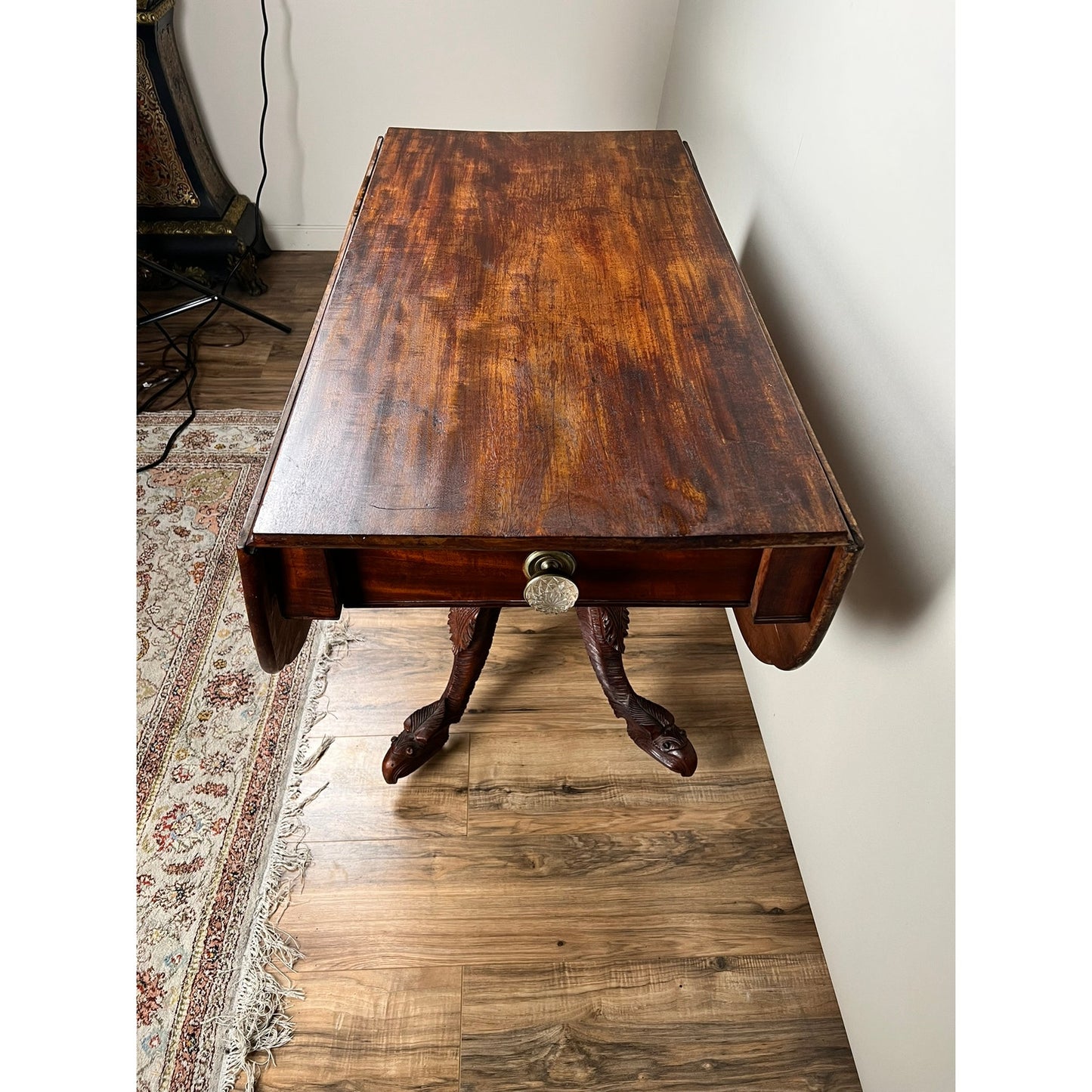 An Antique American Empire Drop Leaf Table with Great Eagle Carved Feet c. 1840 stands on a hardwood floor beside a patterned rug and a white wall.