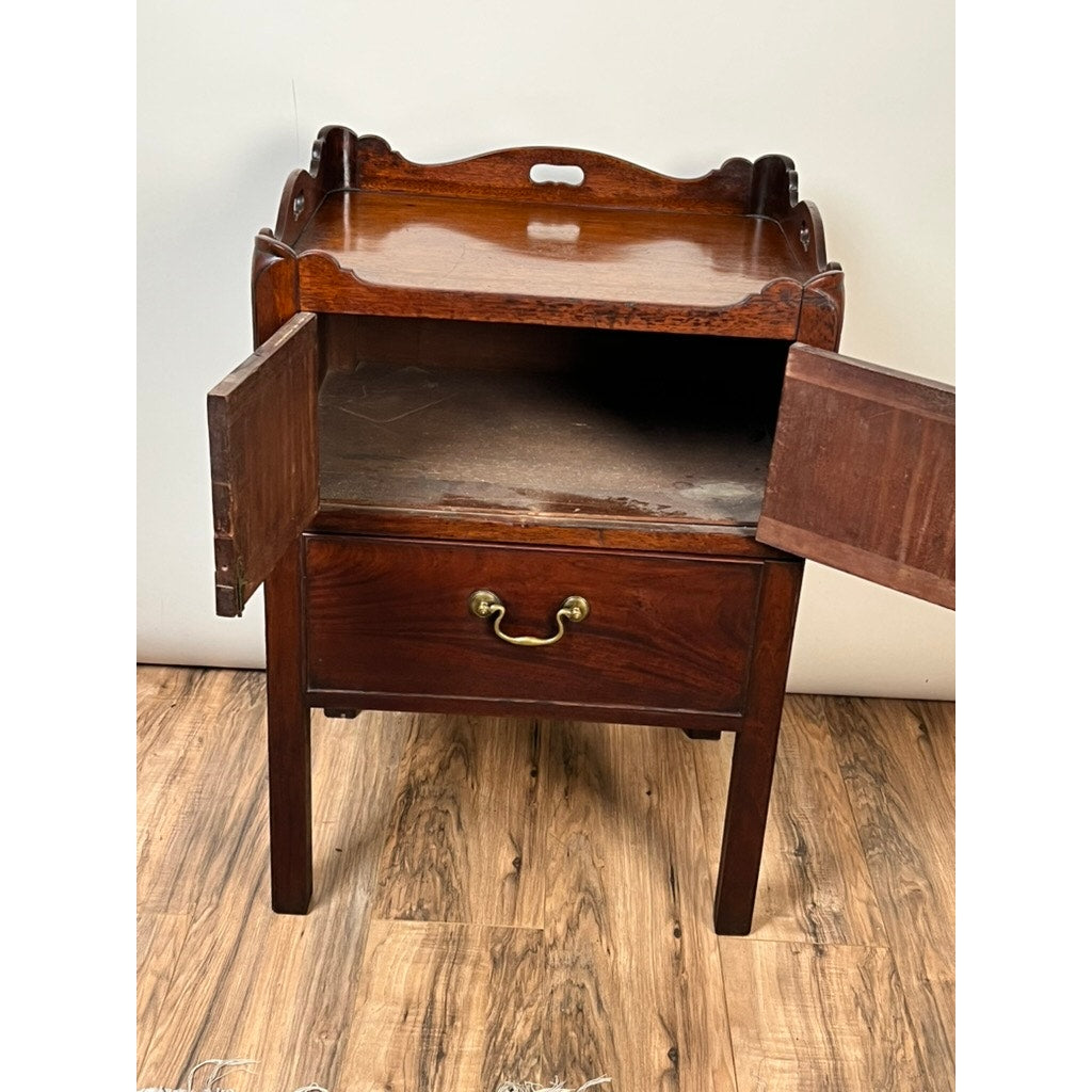 An Antique George III Bedside Commode with pull out leather surface (late 18th century) stands on a wood floor by a plain wall, its decorative top, open cabinet doors, and closed drawer with brass handle on display.