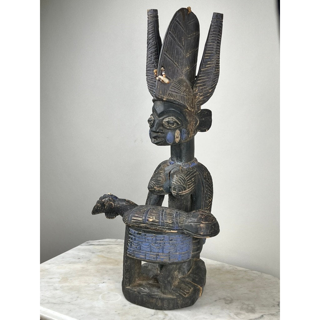 An Antique Igbo Ikenga Shrine Figure, probably from Nigeria, features a seated figure with an elaborate headdress and blue accents holding a drum. Displayed on a light surface against a plain background, it exemplifies exquisite Igbo artistry.