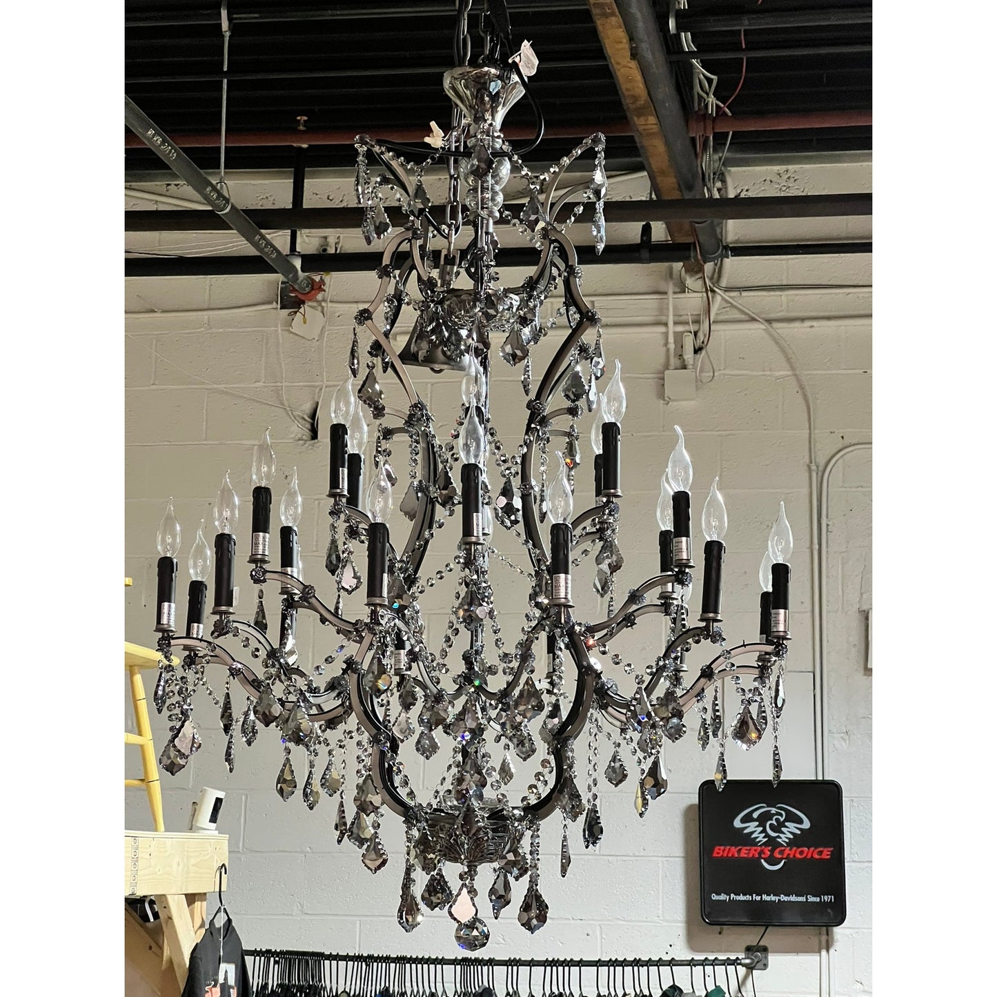 A Large Vintage Smoke Crystal 24 Light Chandelier with candle-style bulbs and crystal accents hangs from an industrial ceiling with exposed pipes and a Bikers Choice sign on the wall.
