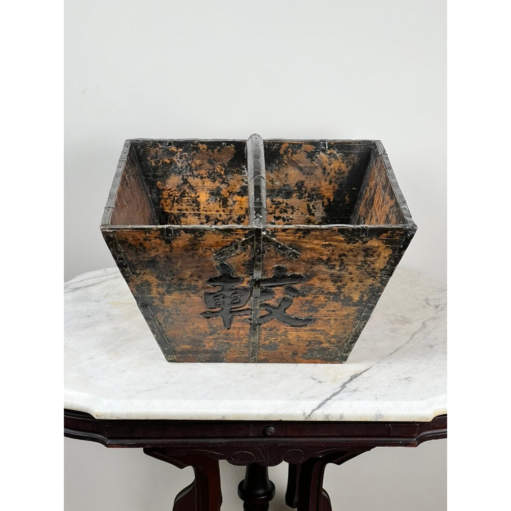 An Antique Chinese Elm Dou Rice Measure, likely from Shanxi, mid-19th century, with a weathered brown-black finish and black Chinese character, sits on a marble-topped dark wood table against a light wall.