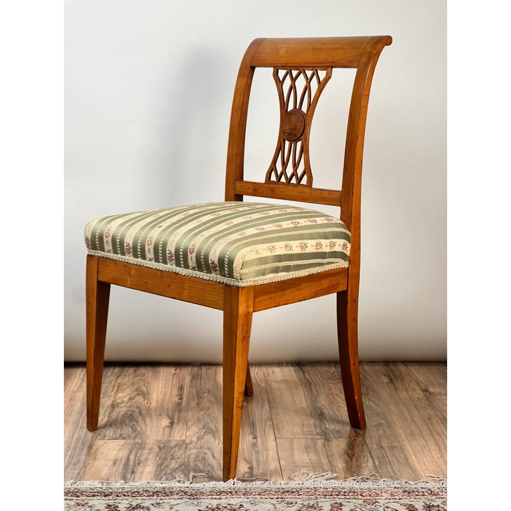 An Antique German Biedermeier Style Cherry Side Chair (c. Early 20th Century) with a carved backrest and green-beige striped cushioned seat sits on a wood floor near a patterned rug, set against a plain light background.