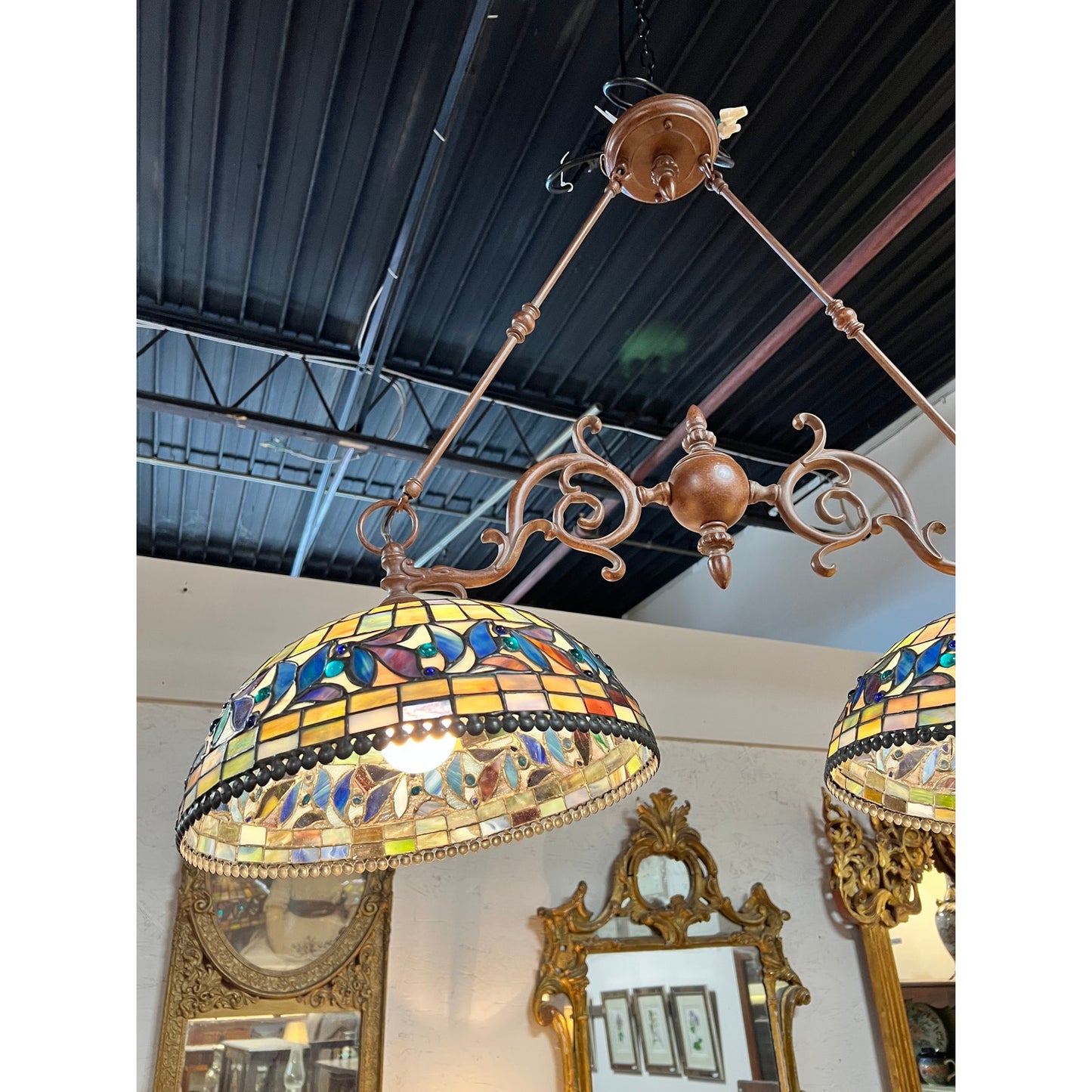 A Vintage Tiffany Style Leaded Stained Glass Double Pendant Billiard Island Light hangs from a black ceiling in a room adorned with antique mirrors and framed artwork on the wall.