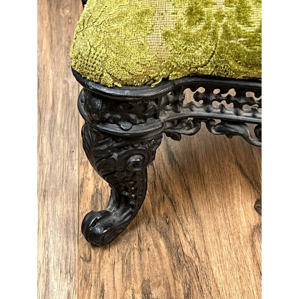 Close-up of an Antique Victorian Cast Iron Foot Stool c. 1900 featuring ornate cast iron legs with intricate carvings and a claw foot, supporting a seat upholstered in green patterned fabric, set on a wooden floor. Brand: Unknown.