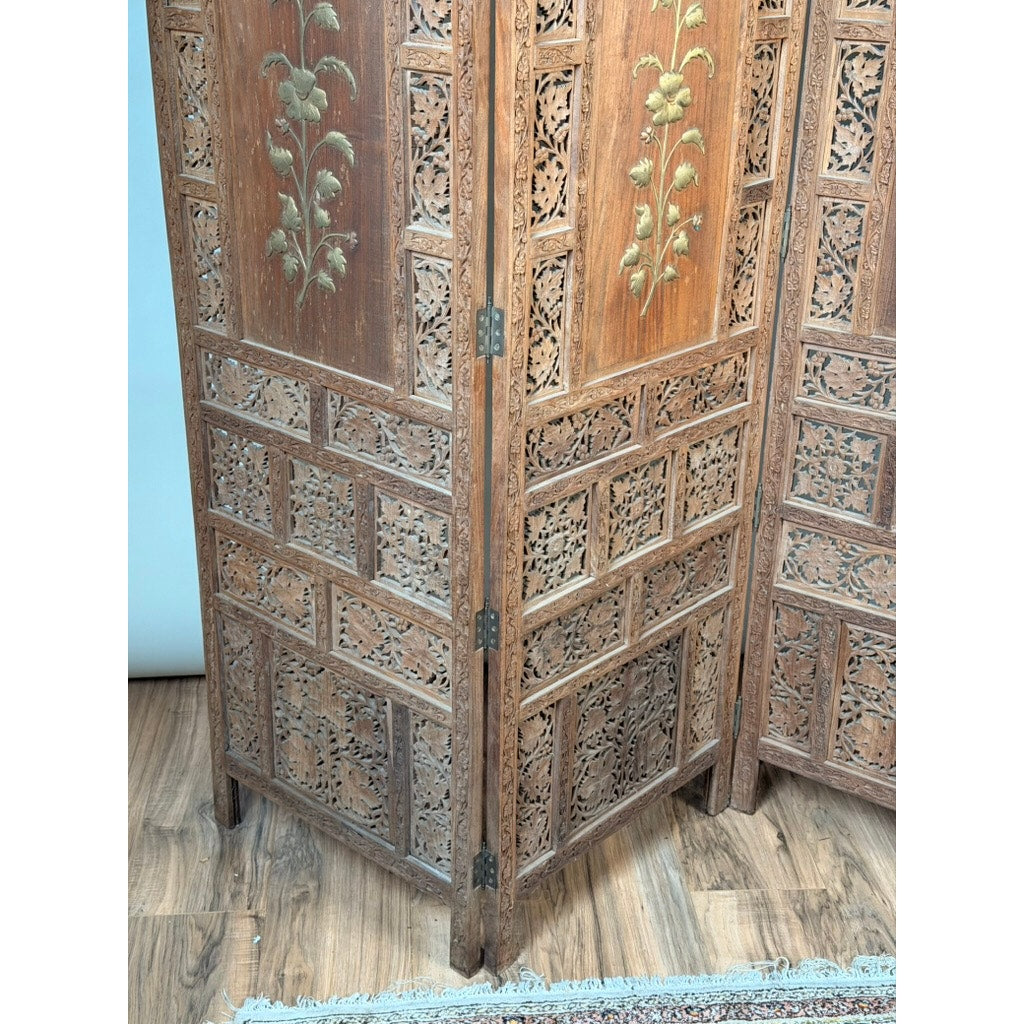 An Early 20th Century Anglo-Indian Carved Teak Four-Panel Screen with applied brass floral decoration by Pearl City Antiques stands gracefully on a wooden floor next to a rug, exemplifying the elegance of an antique decorative screen.