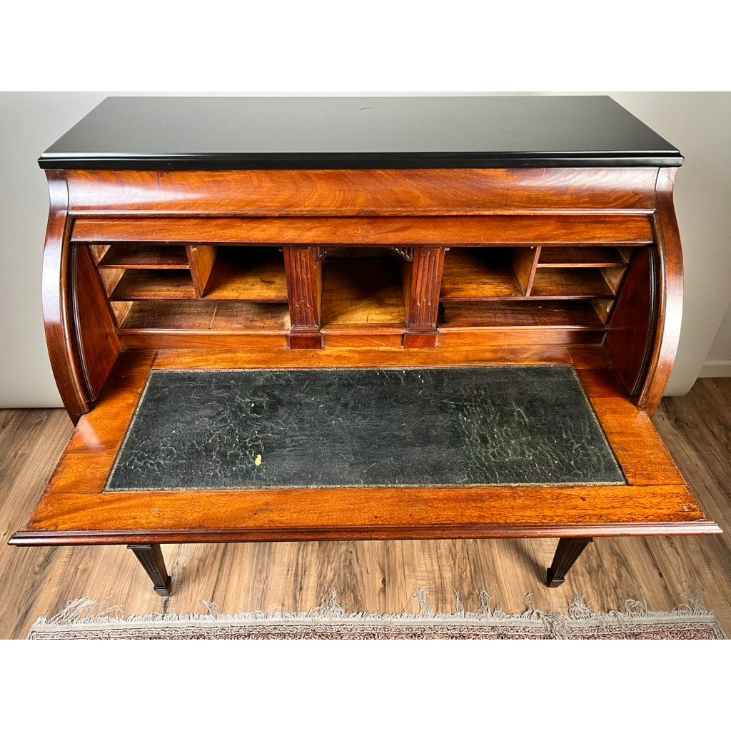 An Antique Directoire Style French Cylinder Desk with Slate Top, dating from the late 19th century, stands on wooden legs over hardwood floors with part of a rug visible, evoking vintage elegance with cubbyholes and upper shelves.