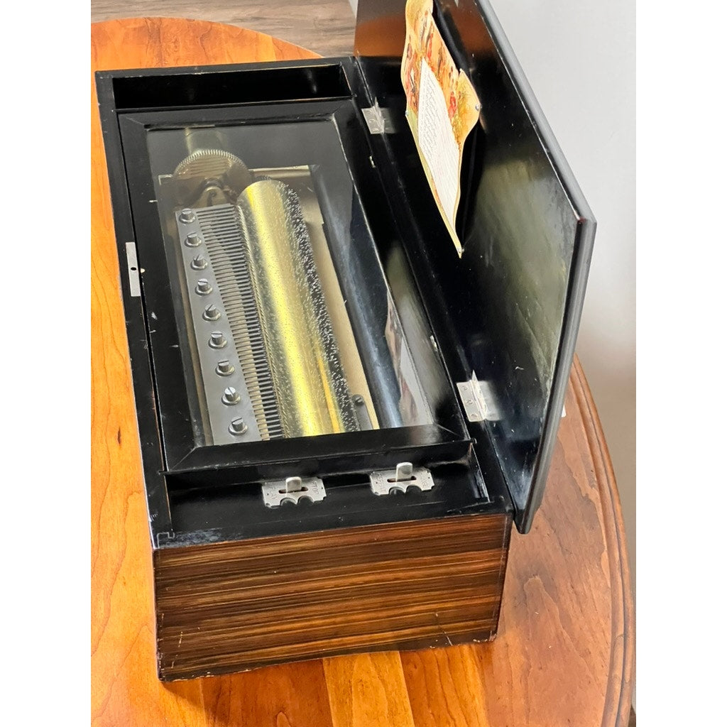 An Antique Swiss 12 Tune Music Box from the late 19th century, with its lid open to show the brass cylinder and metal comb inside its wooden case, sits atop a wooden table. Brand: None/Unknown.