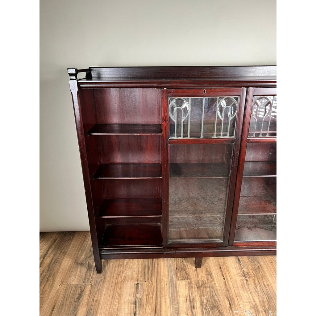 An Antique Arts & Crafts Leaded Glass Mahogany Bookcase c. 1920 with open shelves on the left and two stained leaded glass doors at the top right stands on a wood floor against a light-colored wall.