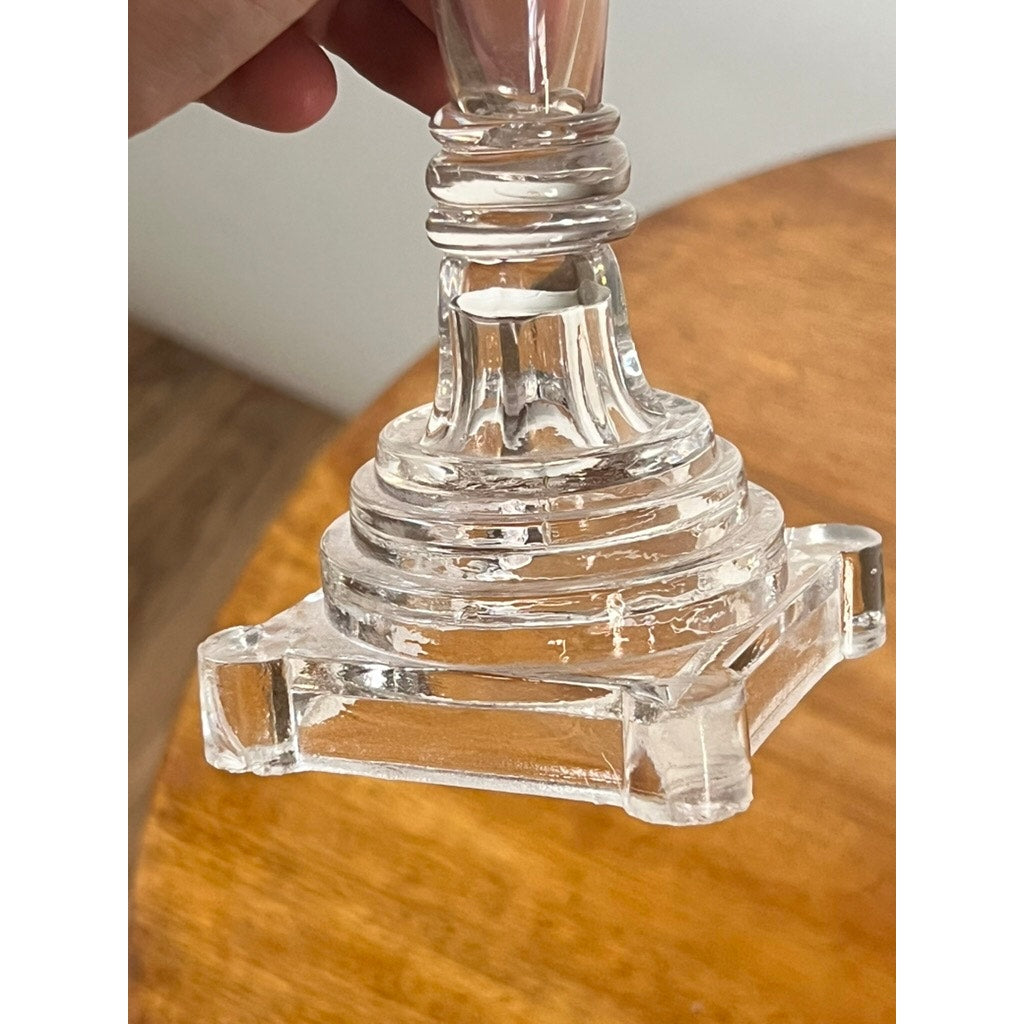 A close-up of an Attributed Boston & Sandwich Pressed Glass Boudoir Fluid Lamp, circa 1850, its clear, tiered square base held above a wooden table. Reminiscent of early American lighting, the elegant form recalls the craftsmanship associated with historic Boston & Sandwich Glass Company pieces.