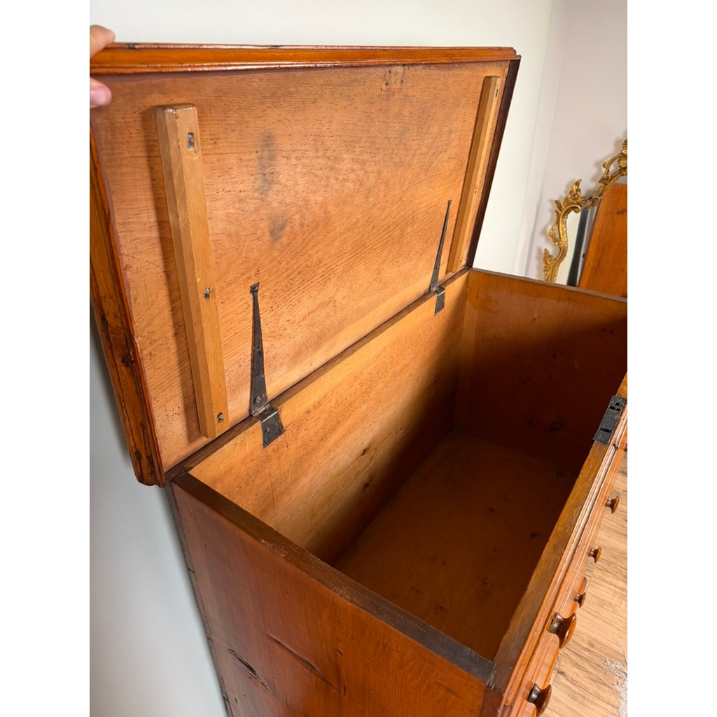 Open wooden chest with hinged lid reveals an empty interior. Crafted in the tradition of early American country furniture, this Late 18th Century New England Pine Two-Drawer Blanket Chest, c. 1780–1810 from Pearl City Antiques features sturdy metal hinges and interior wooden supports beneath the lid. A decorative gold-framed mirror is partially visible in the background, enhancing its period charm.