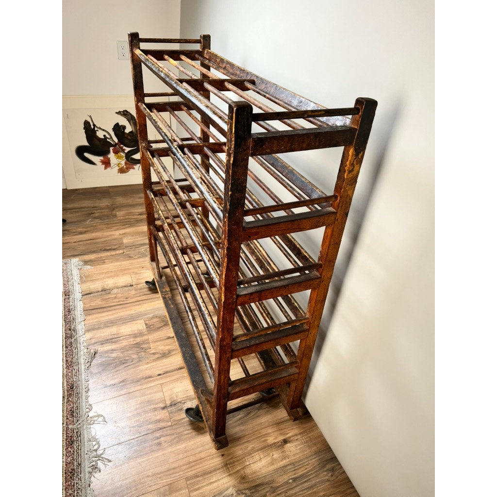 An Antique Oak Shoe Factory Rolling Rack on original industrial casters (late 19th c.) stands against a white wall on a wood floor, featuring a dark, worn finish, open slats, and multiple shelves. A rug and wall art are partially visible nearby.