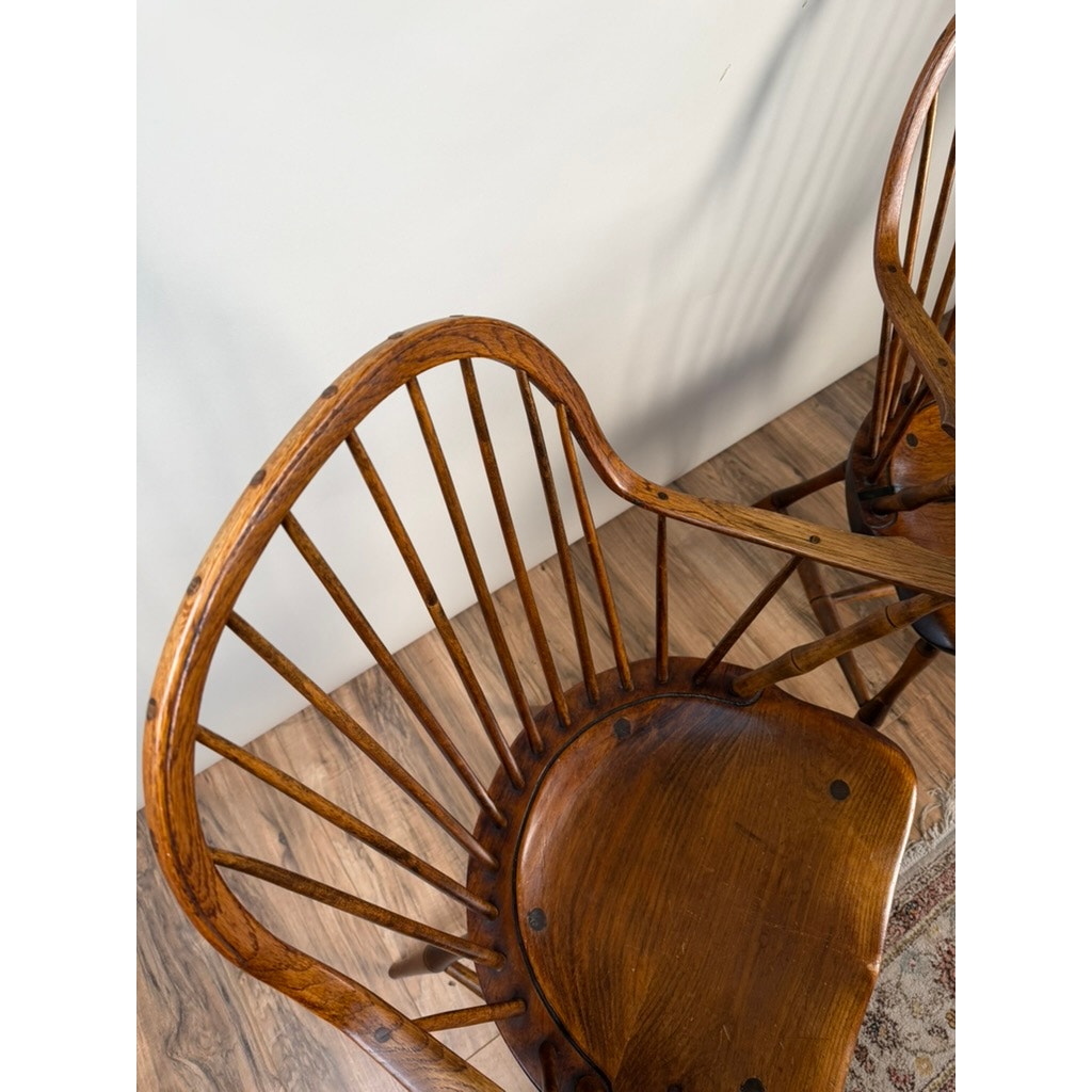 A pair of signed L.E. Partridge Connecticut bench-made continuous-arm Windsor chairs from the 1960s, featuring curved spindle backs and contoured wooden seats, sit on a wooden floor beside a patterned rug, casting gentle shadows on a white wall and evoking the charm of early American craftsmanship.
