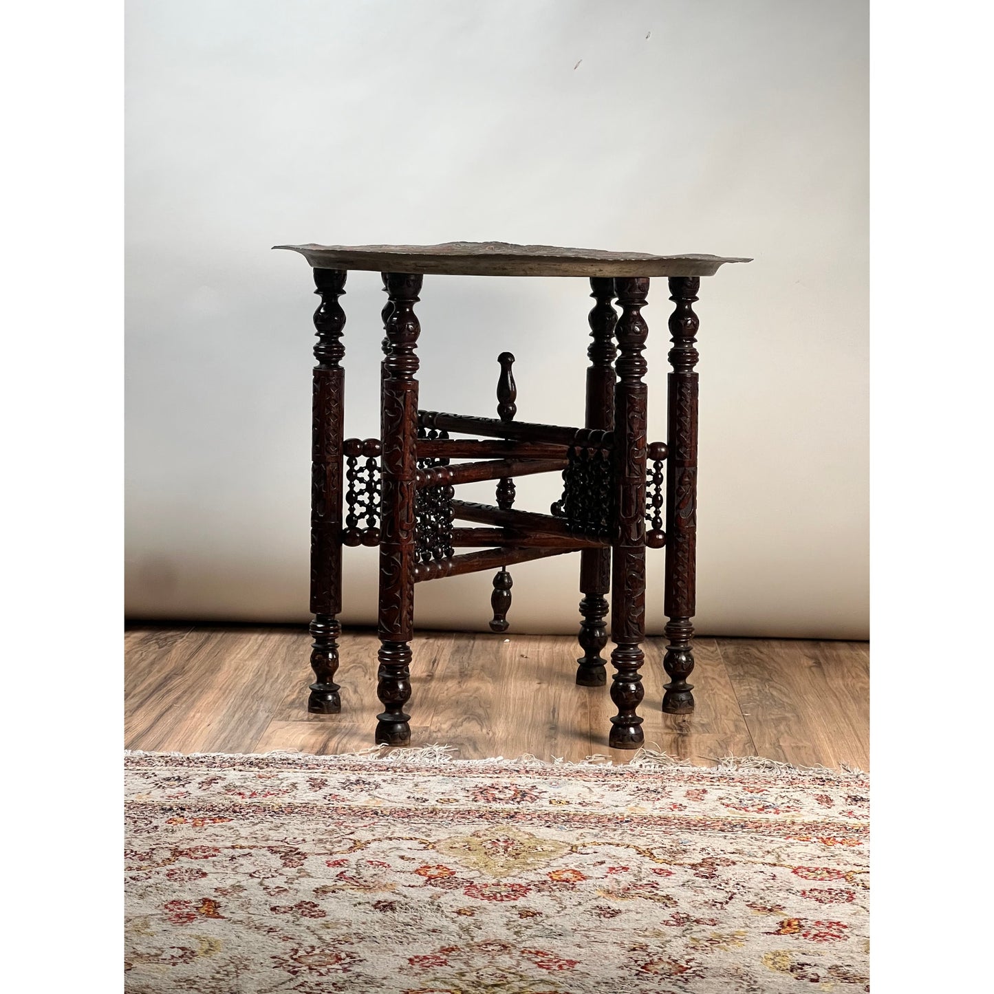 Moroccan or Syrian Moorish folding tray table with intricately carved legs and stretcher, displayed on a wooden floor next to a patterned rug, set against a plain light background.