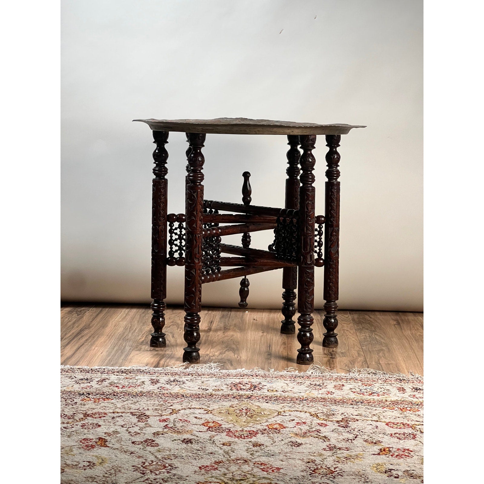 Moroccan or Syrian Moorish folding tray table with intricately carved legs and stretcher, displayed on a wooden floor next to a patterned rug, set against a plain light background.