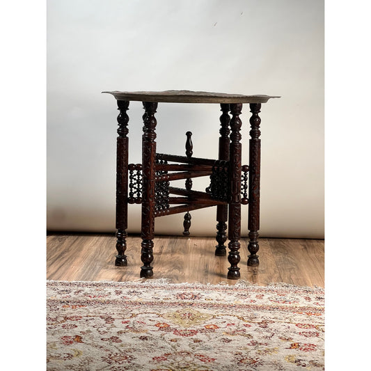 Moroccan or Syrian Moorish folding tray table with intricately carved legs and stretcher, displayed on a wooden floor next to a patterned rug, set against a plain light background.