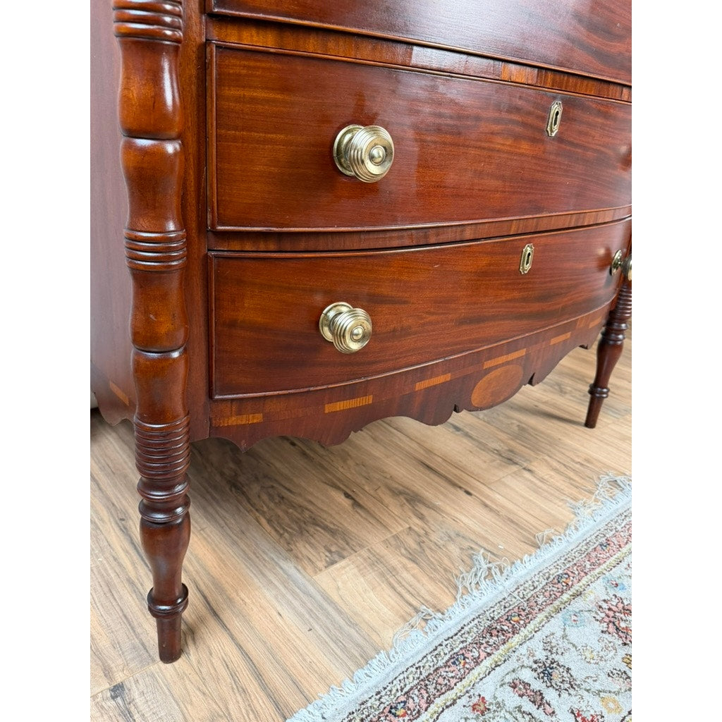 Close-up of an Early 19th Century New England Federal Mahogany Bow-Front Chest of Drawers with Backsplash, c. 1820–1840, featuring brass knobs, decorative inlays, turned legs, and a scalloped apron. This elegant chest stands on a light wood floor next to a patterned rug and exemplifies the refined style of the Federal period.