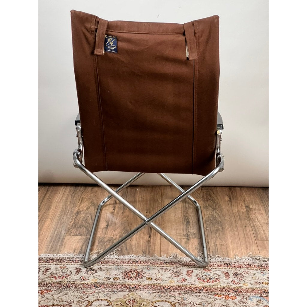 A Vintage Uchida Z Chair Japanese Modern Chrome Sling Lounge Chair with Headrest by Uchida, featuring a brown sling and chrome metal frame, is positioned with its back facing the camera as it rests on a wooden floor and partially on a patterned rug.