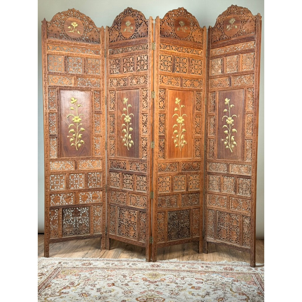 Early 20th Century Anglo-Indian Carved Teak Four-Panel Screen with Applied Brass Floral Decoration by Pearl City Antiques, showcasing intricate woodwork and elegant brass floral accents. Set upon a hardwood floor with a patterned rug in front, this antique screen serves as a sophisticated room divider, bringing timeless charm and artistry to any interior.
