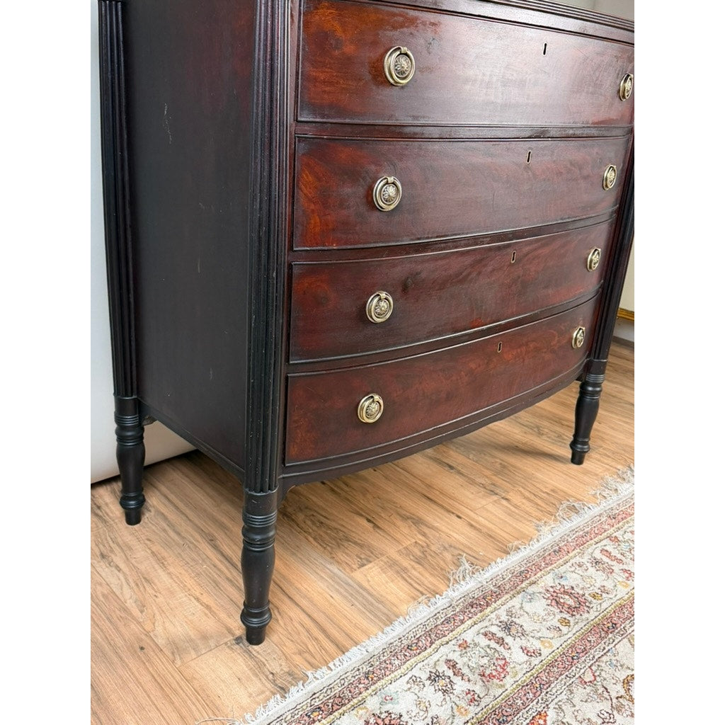 An Early 19th Century New England Federal Mahogany Bow-Front Chest of Drawers (c. 1810–1825), featuring four curved drawers and round metal handles, stands on a wooden floor beside a patterned rug.