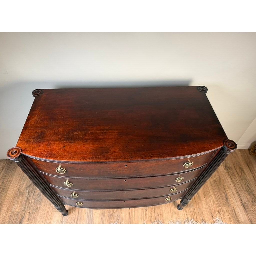 An early 19th-century New England Federal mahogany bow-front chest of drawers, dating from circa 1810–1825, features four curved drawers with round metal pulls and decorative side columns. It stands on a wooden floor against a plain wall.