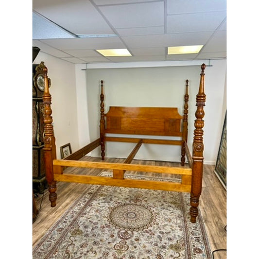 A Pearl City Antiques “Antique 19th Century Sheraton Field Bed Resized to King by Leonards New England Seekonk, MA” with finely carved posts stands on a patterned rug in a room featuring wood flooring, white walls, and a grandfather clock in the corner. Fluorescent lights are fitted into the ceiling.