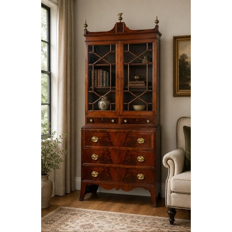 A Federal Flame Mahogany Secretary Bookcase with Eagle Finial, dating from circa 1810–1825, featuring glass doors on top, brass handles, and four drawers, stands beside a beige sofa near a window and a framed painting. A potted plant sits on the floor to the left.