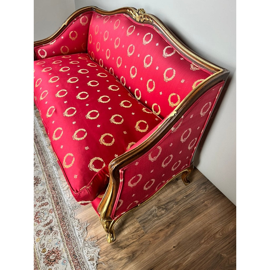 A Vintage French Regency Style Custom Upholstered Settee with a giltwood frame and red fabric featuring gold accents and a circular pattern sits against a white wall on a wooden floor next to a patterned rug. Brand: Unknown.