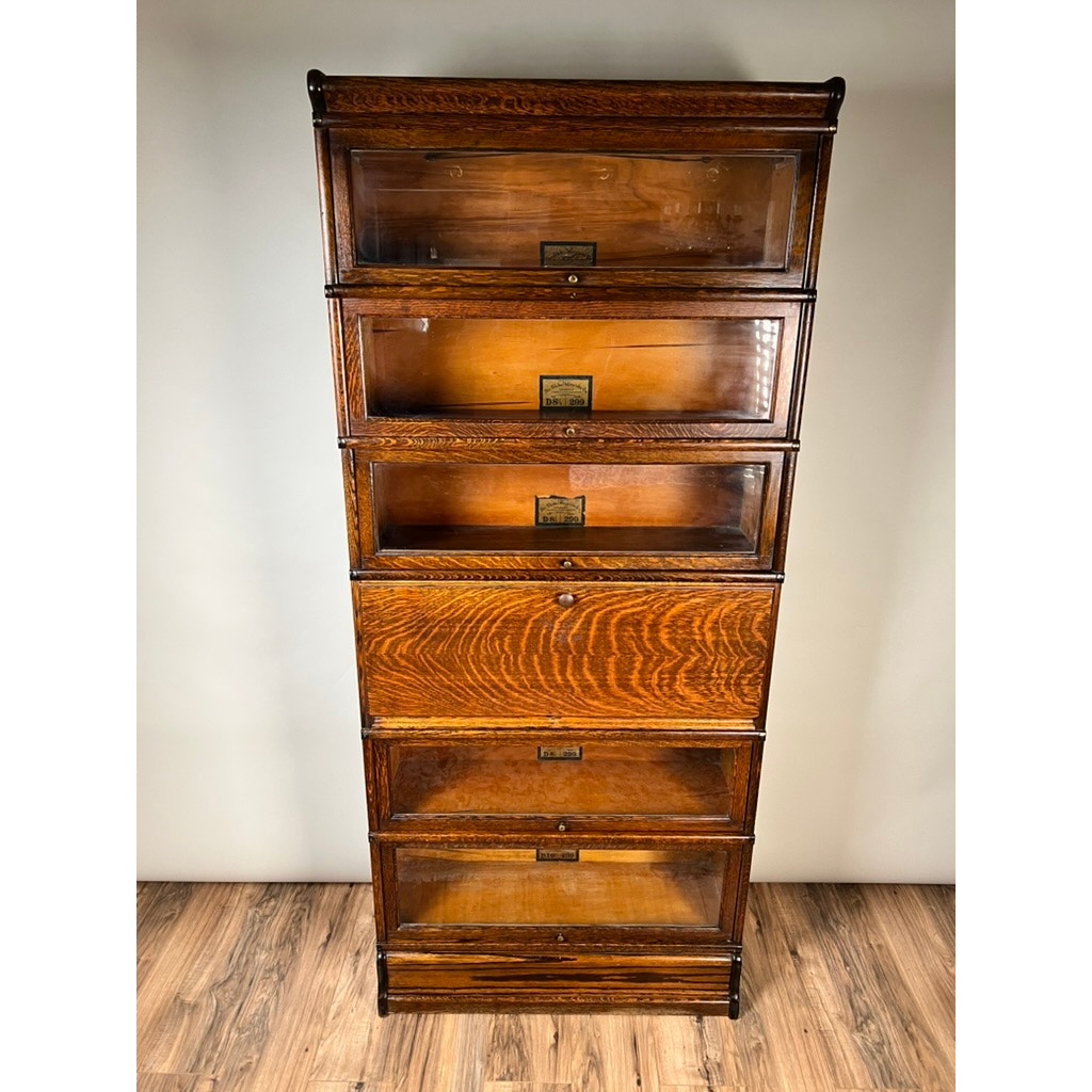 Standing against a plain wall on a wooden floor, this impressive Antique Globe-Wernicke Quarter Sawn Oak 6-Stack Barrister Bookcase Secretary Desk (c. 1905–1915) by Globe Wernicke features six stacked glass-front sections with brass label holders and a lustrous, polished finish in quarter-sawn oak.