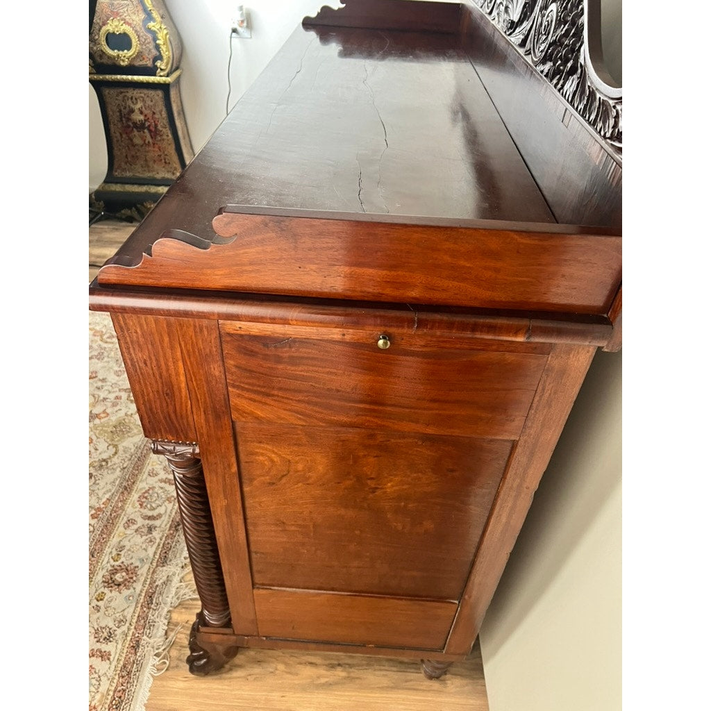 A close-up of an Antique American Empire Mahogany Sideboard with rosewood edge banding, c.1840, probably from Philadelphia, displays carved details and a keyhole on its side panel. It stands on hardwood flooring beside a patterned rug and ornate cabinet.